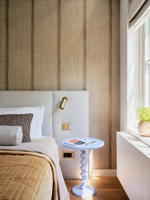 Een slaapkamer met een bed, een blauw bijzettafeltje, een raam met een plant op de vensterbank en een beige muurbekleding.