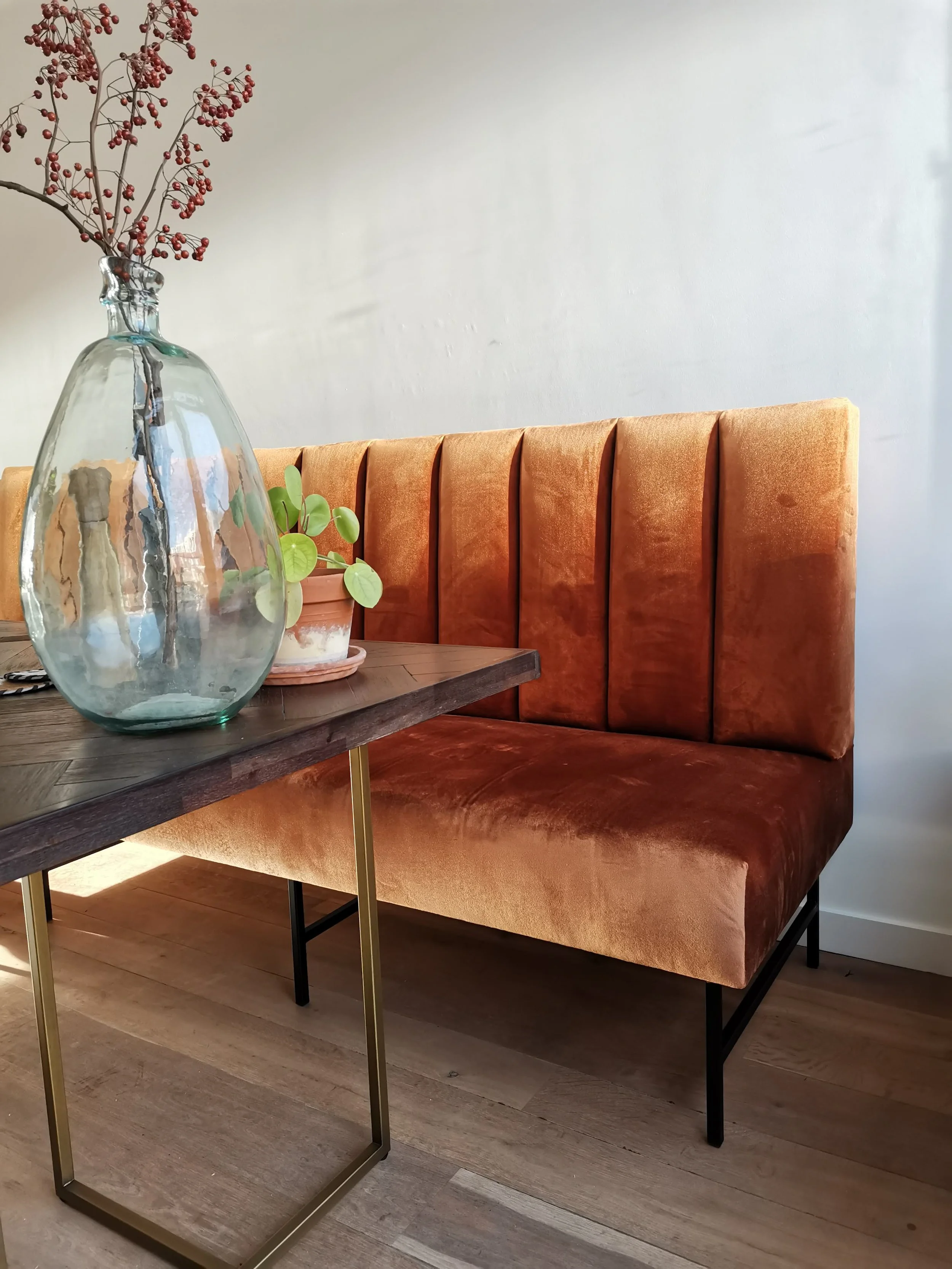 Een glazen vaas met rode bessen en een plant met ronde groene bladeren op een houten tafel, naast een oranje velvet zitbank tegen een witte muur.