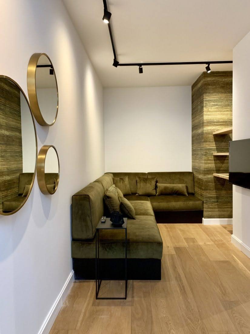 Een woonkamer met een groene bank, een klein bijzettafeltje, drie ronde spiegels aan de witte muur, houten vloer en een muur met houten panelen.