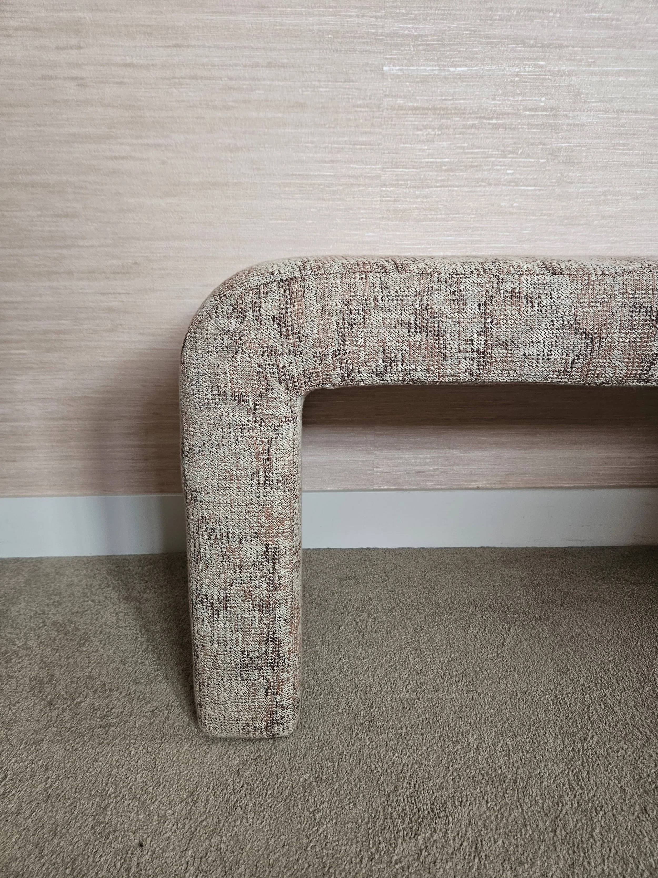 De afbeelding toont een meubelstuk met een tapijten hoes, dat een L-vorm heeft, en staat op een beige tapijtvloer voor een lichtroze en beige gestreepte muur.