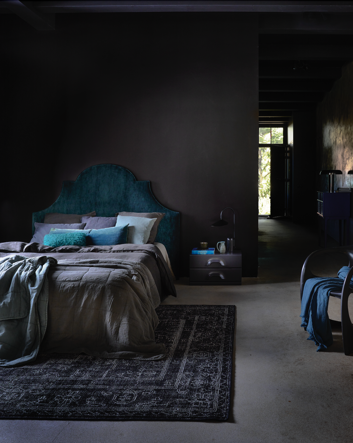 Een slaapkamer met een donker zwarte muur, een bed met een blauwe hoofdbord en diverse kussens, een nachtkastje met decoratie, en een stoel met een blauwe doek, een open deur en een raam met uitzicht op groen buiten.