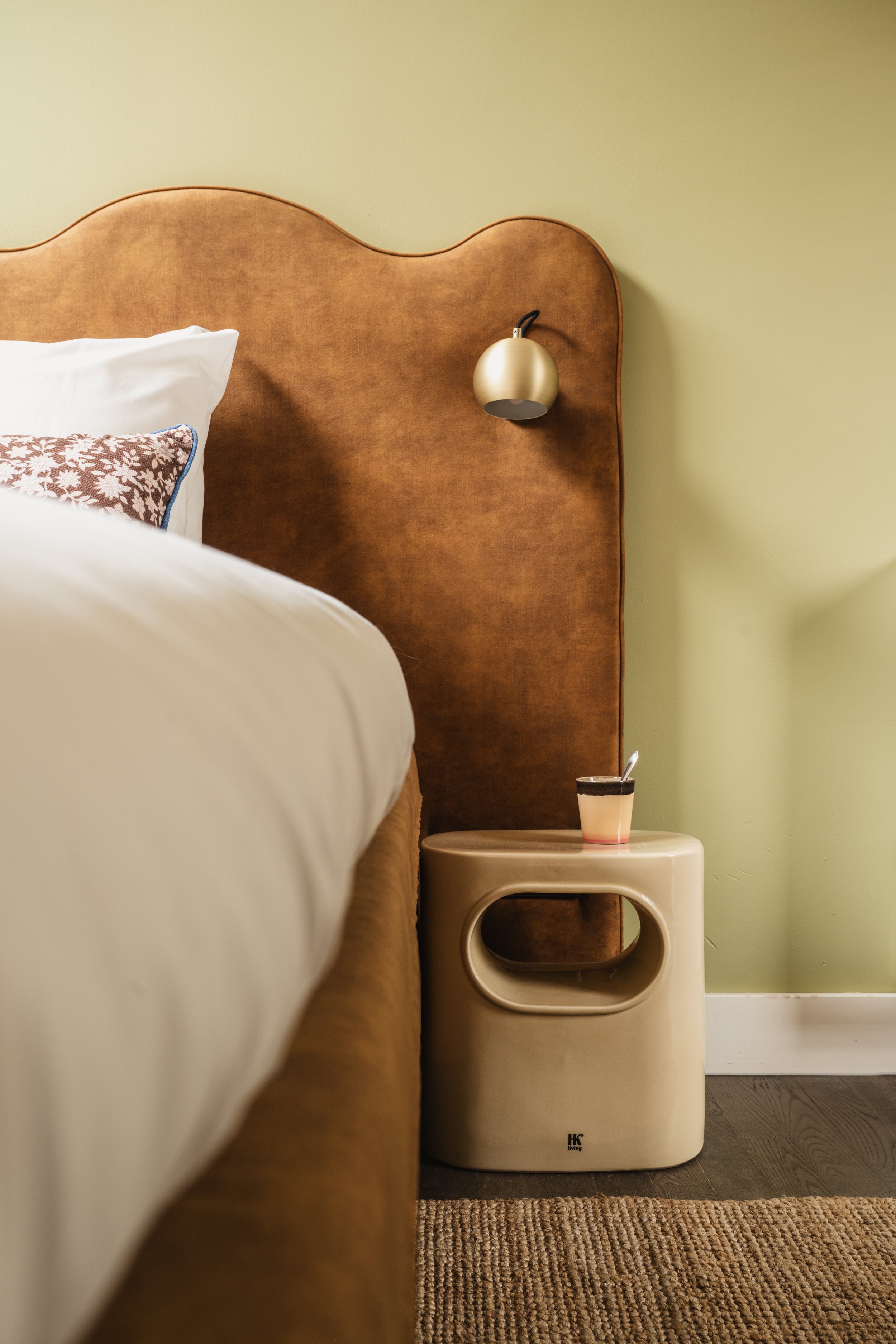 Een slaapkamer met een bruine hoofdbord, een witte bedrand en een kussensloop met een bloemenpatroon. Aan de muur hangt een gouden wandlamp en op een beige nachtkastje staat een glas met een drankje.