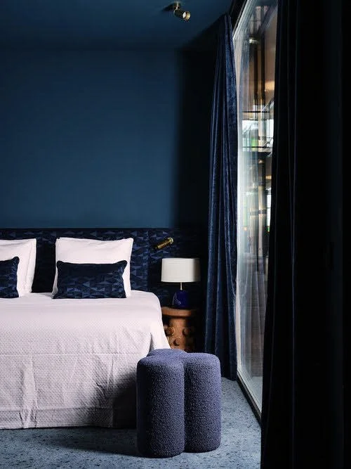 Een slaapkamer met een wit bed, blauwe kussen en beddengoed, een nachtkastje met een lamp, blauw gordijn en een groot raam met balkon. 