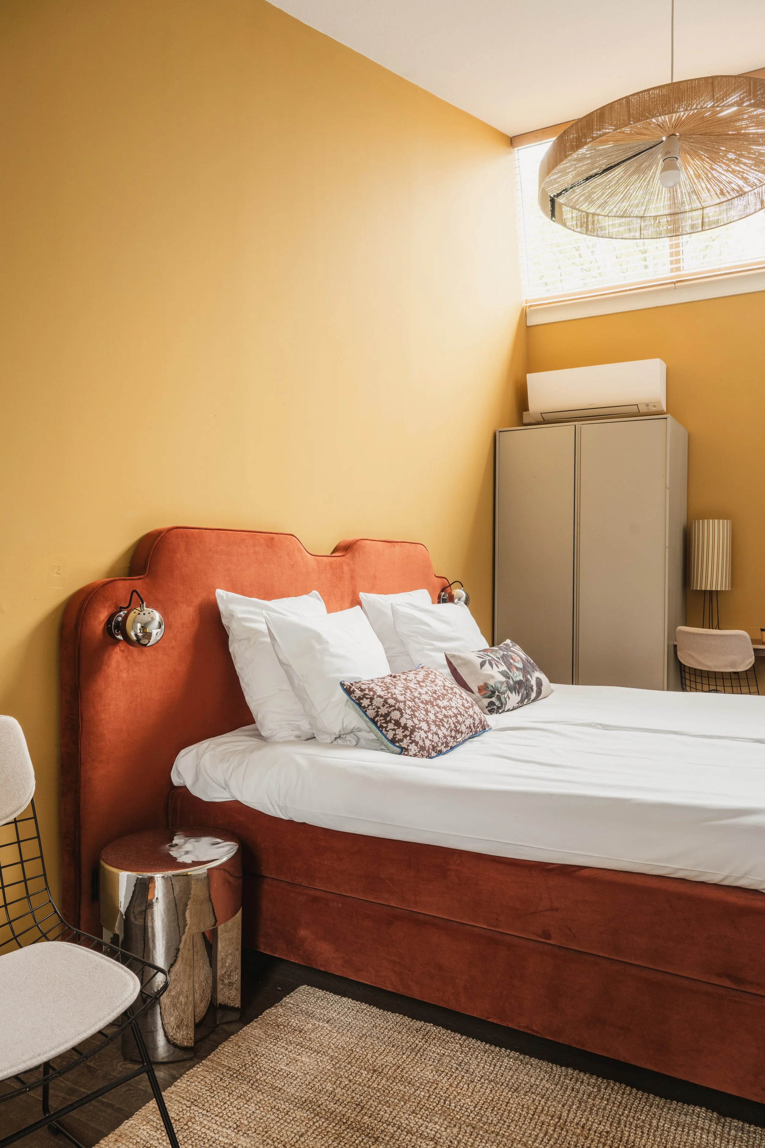 Een slaapkamer met een oranje hoofd- en voeteneinde van het bed, witte lakens en verschillende kussens, een nachtkastje met een spiegel en een stoel.