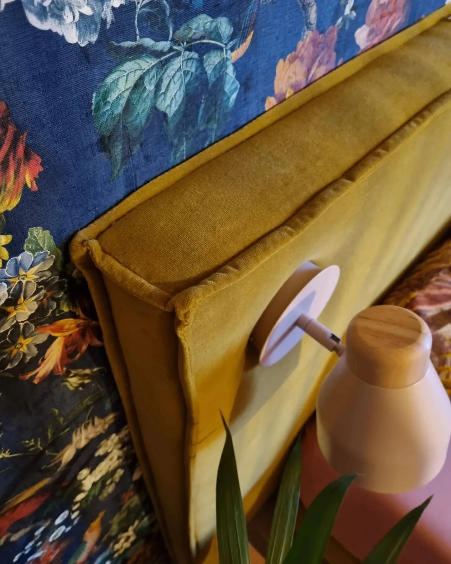 Close-up van een kleurrijk bedding met bloemen, naast een gele bekleding van een meubelstuk en een houten lamp op een tafeltje.