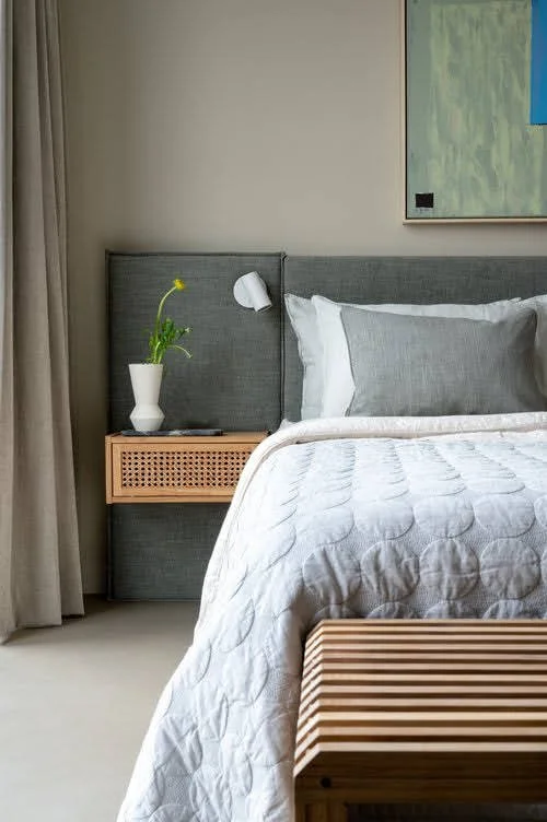 Een bed met witte en grijze lakens, een nachtkastje met een witte vaas en een groene plant, en een abstract schilderij aan de muur.