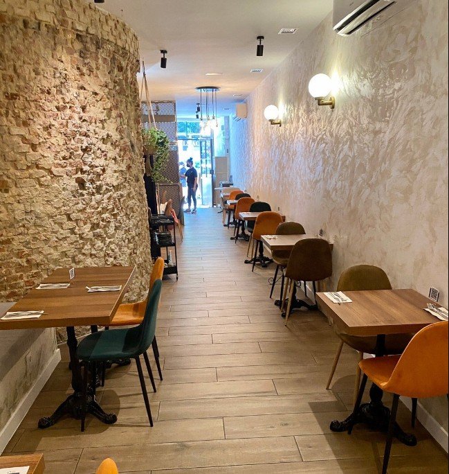 Empty dining area in a restaurant with wooden tables, colorful chairs, and exposed brick wall on the left.