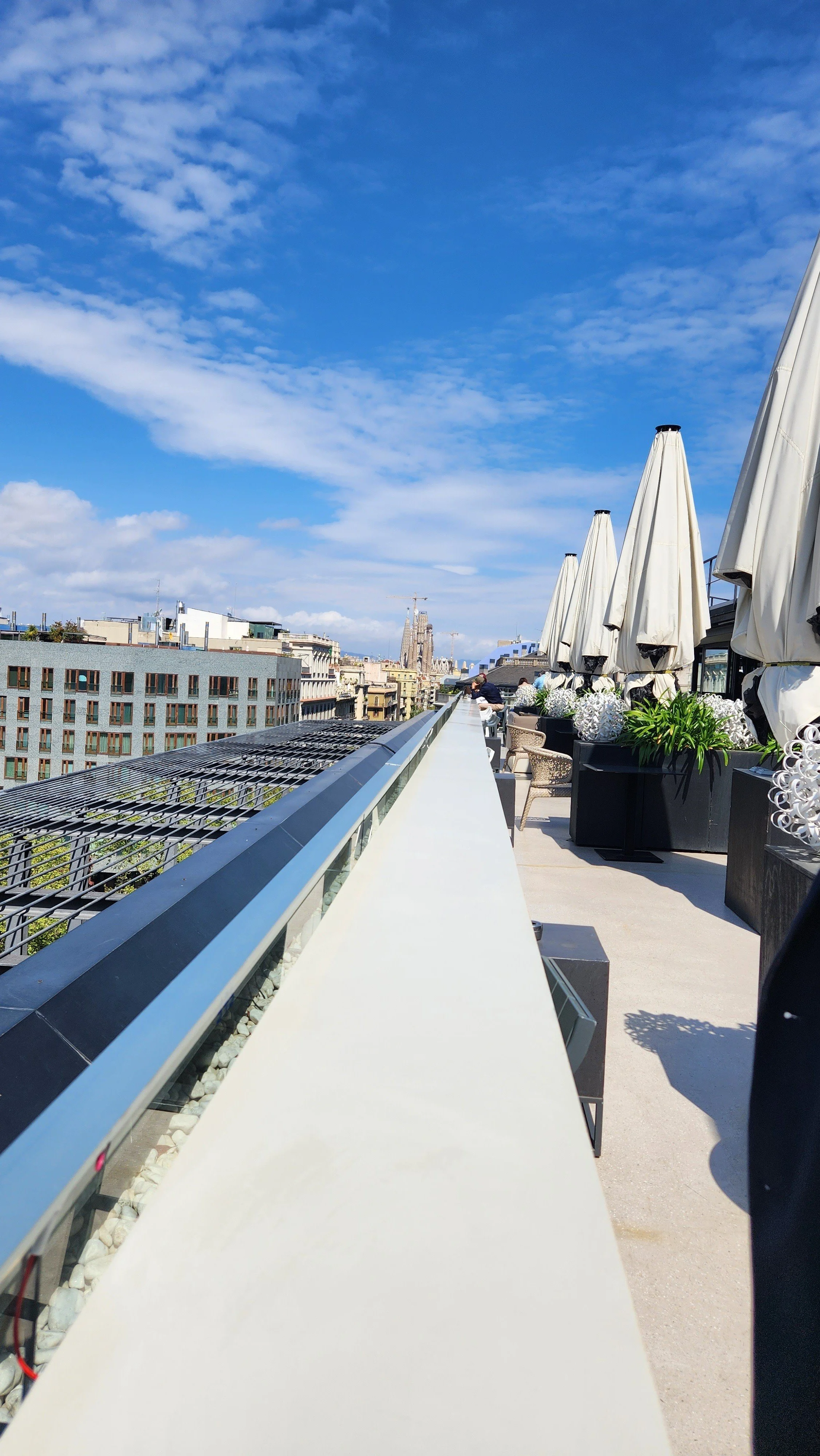 Rooftop terrace with blue sky, white clouds, standing umbrellas, potted plants, and city buildings in the background.
