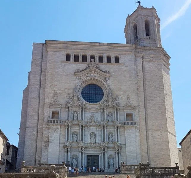 Facade of an old stone church with ornate architectural details and sculptures, featuring a large circular window, a tall bell tower, and a group of people at the entrance, under a clear blue sky.