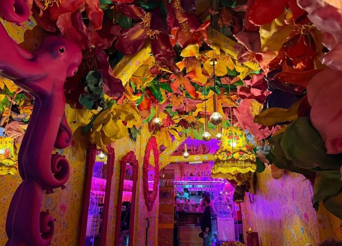 Colorful interior decorated with large artificial flowers on the ceiling, pink and yellow walls, ornate lampshades, and pink neon lighting. A person is seen standing at a counter in the background.