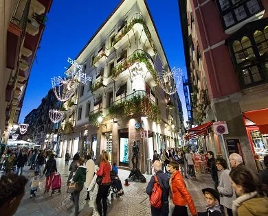 Busy city street at night with a multi-story building decorated with Christmas lights and ornaments, pedestrians walking on the sidewalk.