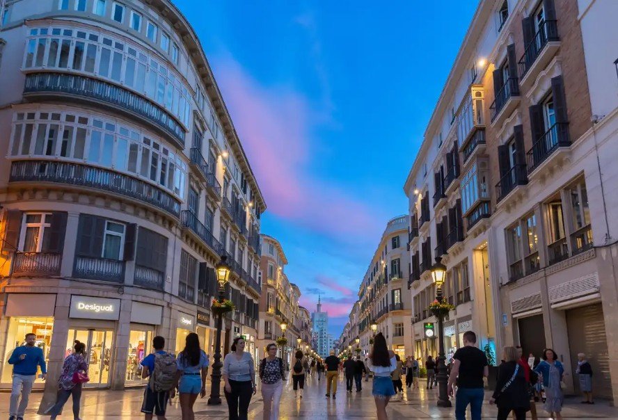 Dusk on a busy shopping street with people walking, luxury stores like Desigual, historic European architecture, decorative street lamps, and a colorful sky.