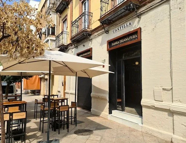 Outdoor patio with tables and chairs shaded by a large umbrella in front of a building with balconies, and signs that read 'Castilla' and 'Barra Triana Puerta'.