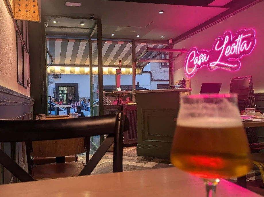 Interior of a restaurant or bar with a neon pink sign that reads 'Para Yeta', a partially visible beer glass in the foreground, and an outdoor seating area visible through large glass windows.