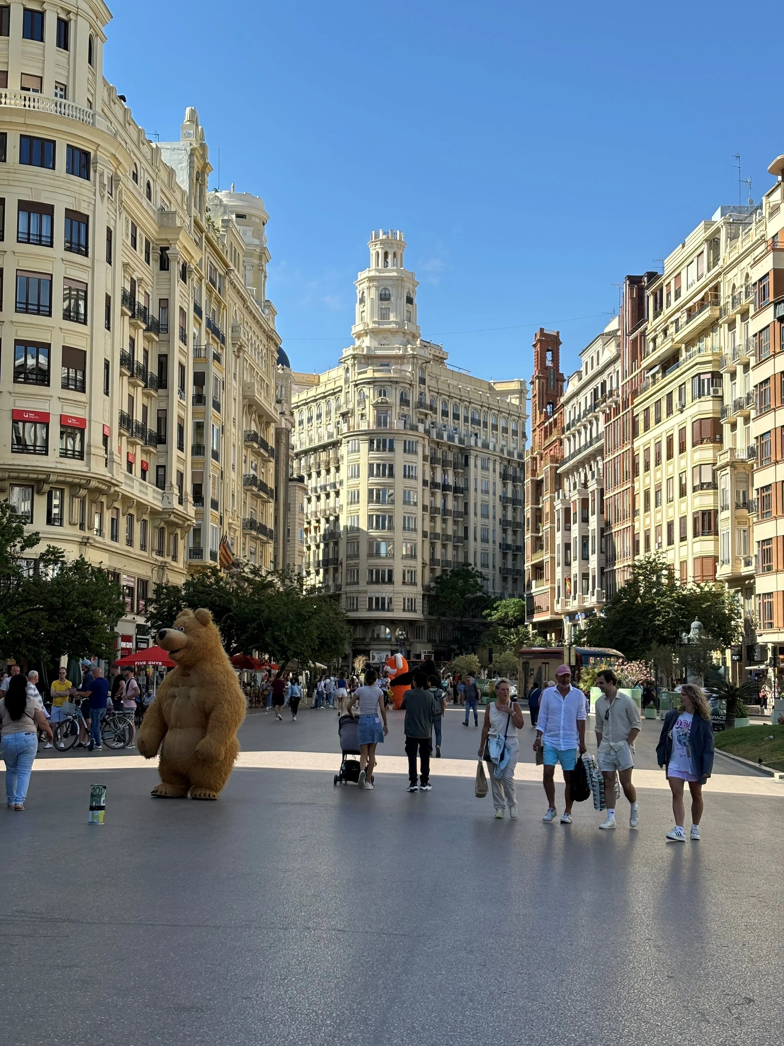 People walking and cycling in a busy city square with tall, ornate buildings and blue sky, featuring a person in a bear costume in the foreground.