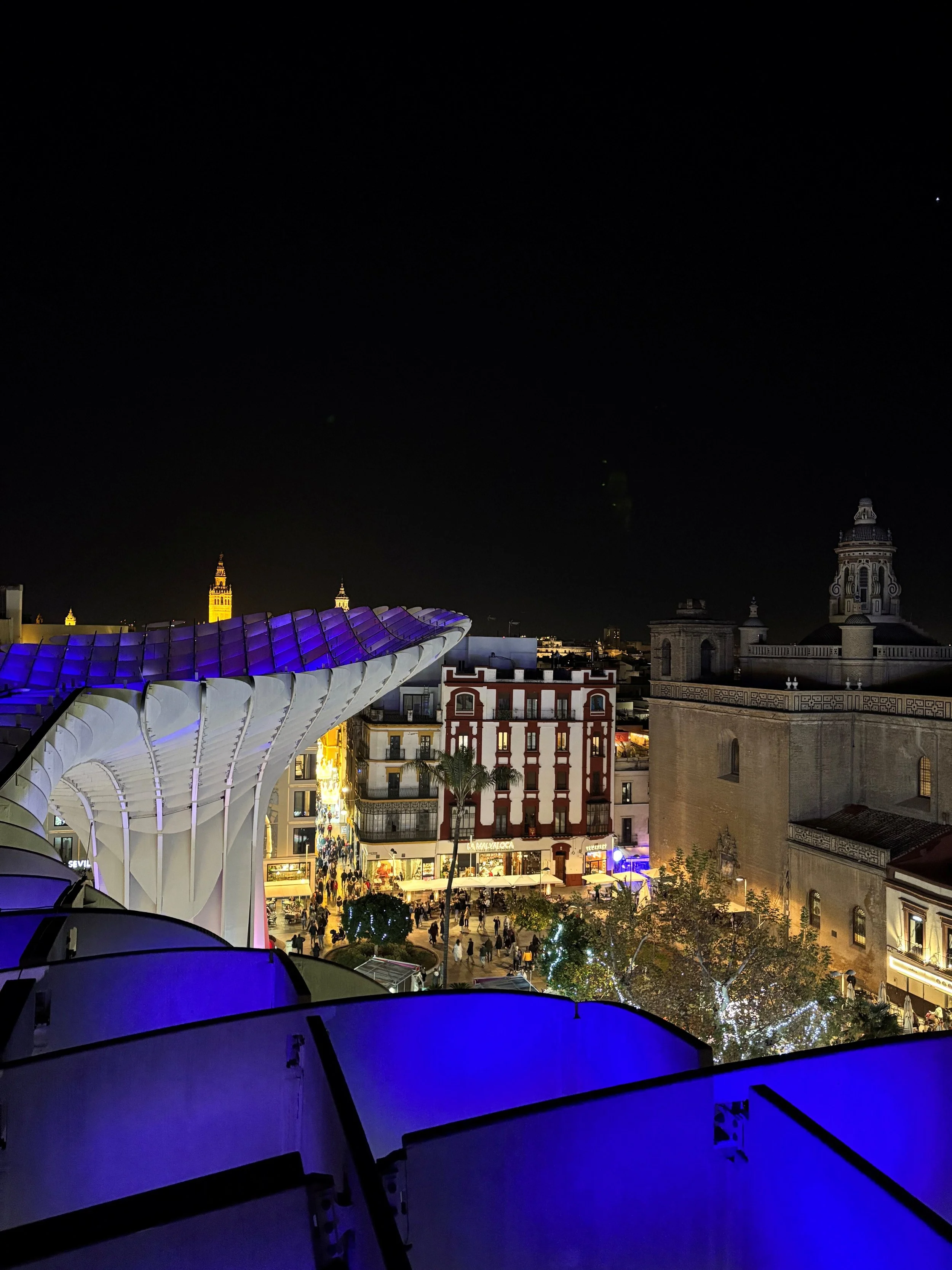Nighttime view of a city with a modern, uniquely shaped stadium/building illuminated in purple, with historic buildings in the background, and a crowd of people in a lively downtown area.