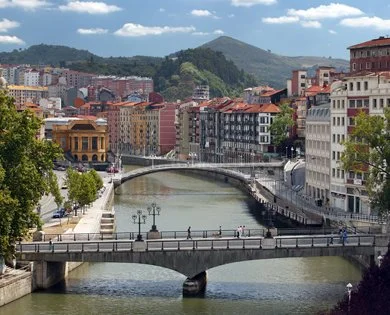 Cityscape with colorful buildings along a river, bridges crossing over, green hills in the background, and a partly cloudy sky.