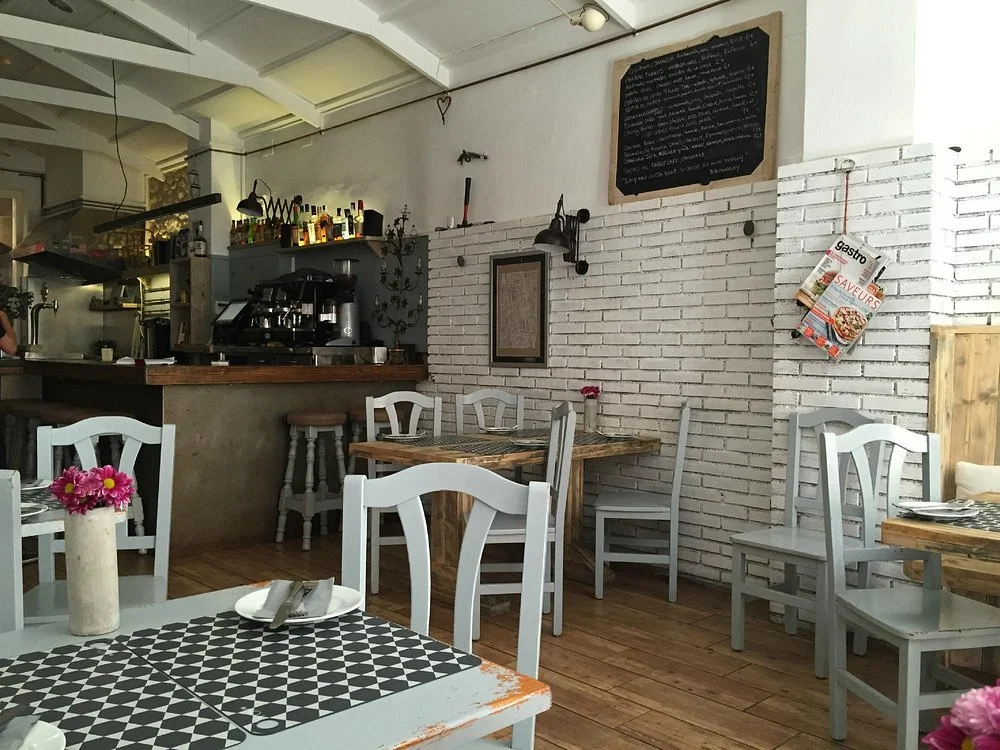 Interior of a cozy cafe with white chairs, a wooden table, a brick wall, and a chalkboard menu. There are flowers in vases on the tables and bottles on a shelf behind the counter.