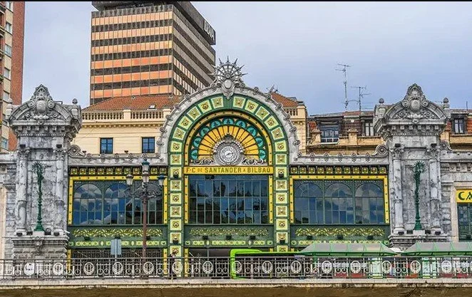 Historic building with ornate architecture, a large clock, and stained glass windows, labeled 'F.C. SANTANDER BILBAO', located on a city street with modern high-rise buildings in the background.