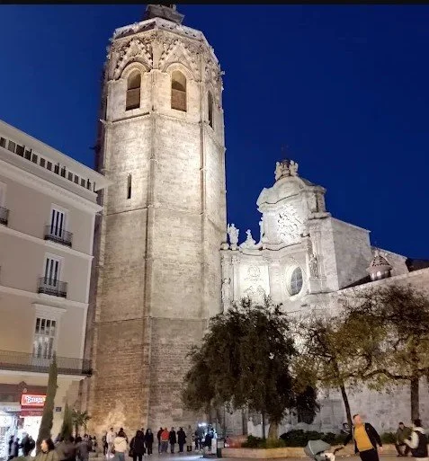Nighttime view of historic stone church with a tall bell tower and ornate facade, surrounded by modern buildings and people walking on the street.
