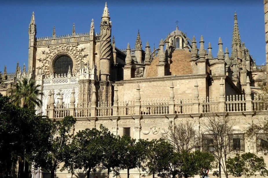 Front view of a large Gothic-style cathedral with intricate stone carvings, spires, and a series of smaller towers, set against a clear blue sky, with trees in the foreground.