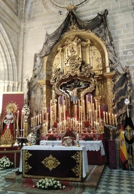 Interior view of a religious altar with numerous lit candles, floral arrangements, gold decorations, and a crucifix, within a church setting.