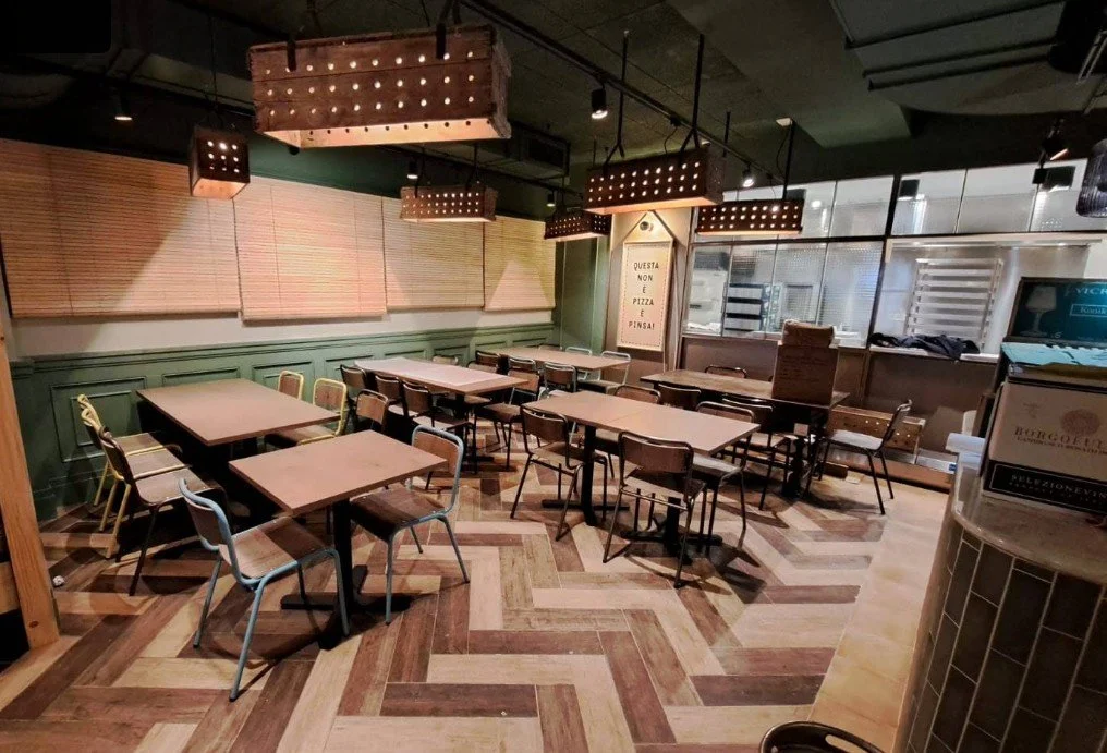 Empty restaurant dining area with wooden tables and mixed chairs, green wainscoting, wooden wall accents, and rectangular hanging light fixtures.
