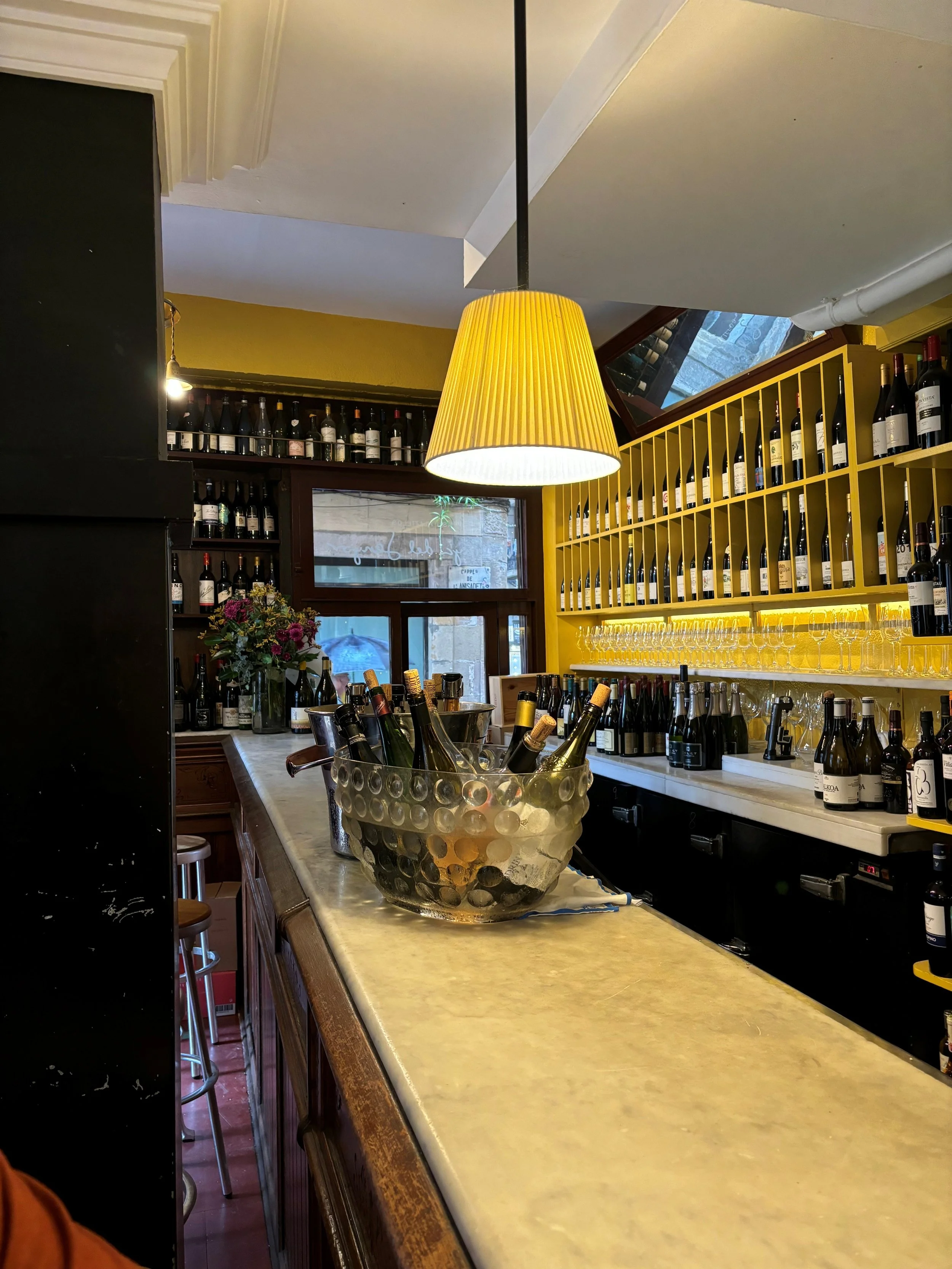 The image shows a cozy wine bar with shelves of wine bottles on the wall, a large yellow hanging lamp, a marble countertop with bottles of wine, glasses hanging underneath, a clear decorative bowl with bottles of wine inside, flowers, and a window in the background.