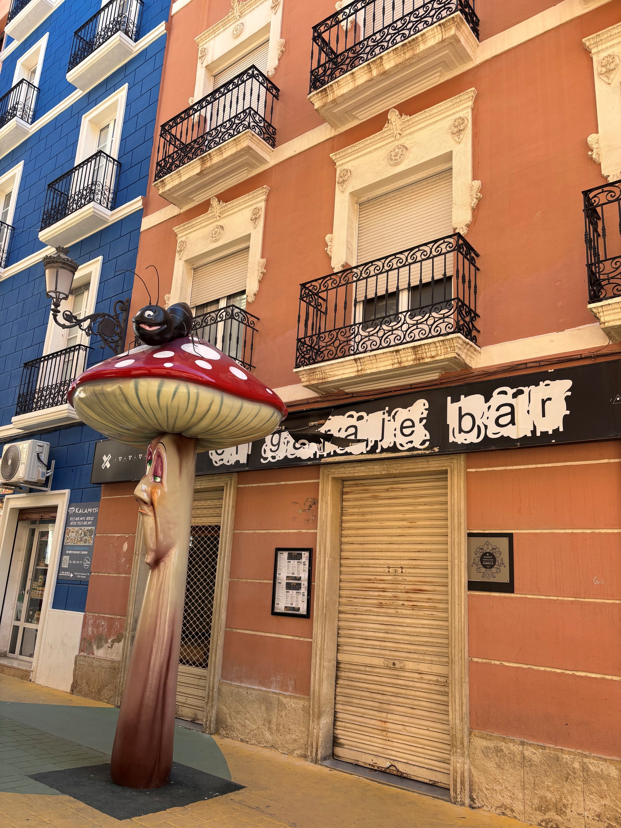 A street scene showing a whimsical sculpture of a mushroom with a smiling, black cartoon-like ant on top. Behind it, colorful buildings with balconies and window shutters, and a storefront with a sign partially visible.