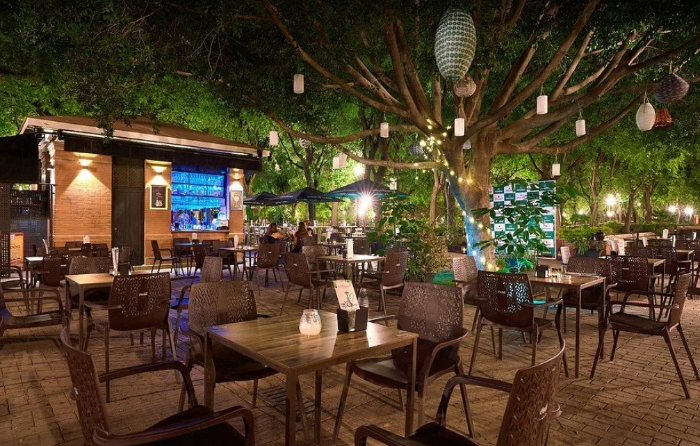 Outdoor dining area at night with tables, chairs, a large tree decorated with hanging lanterns, and a bar with blue lighting in the background.