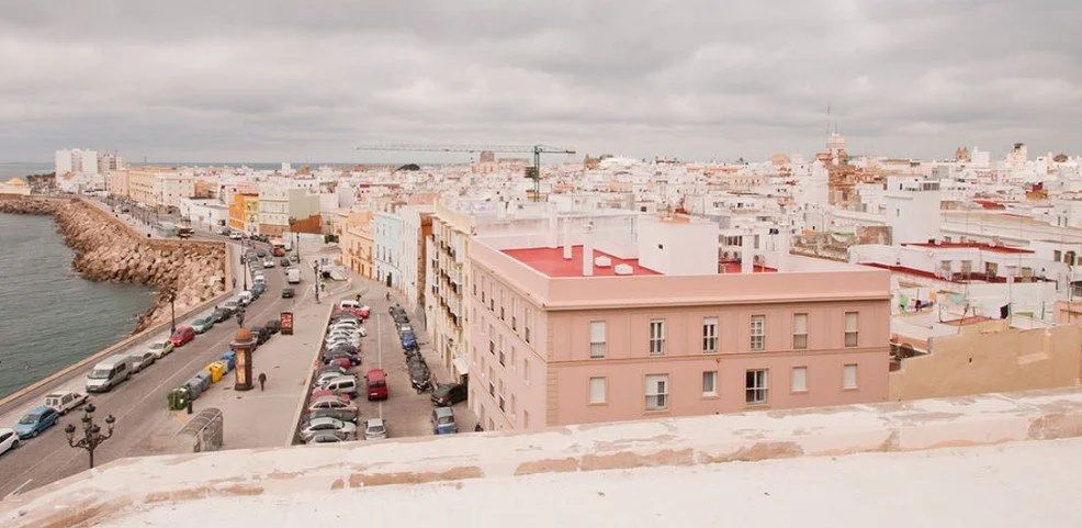 Cityscape with colorful buildings, a street with parked cars, and a waterfront with rocks under a cloudy sky.