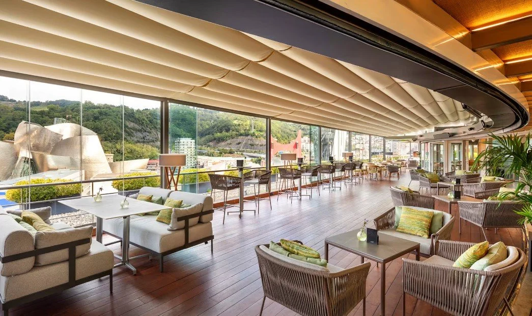 An elegant lounge with large windows overlooking city and greenery, furnished with sofas, armchairs, and small tables, in a contemporary style.
