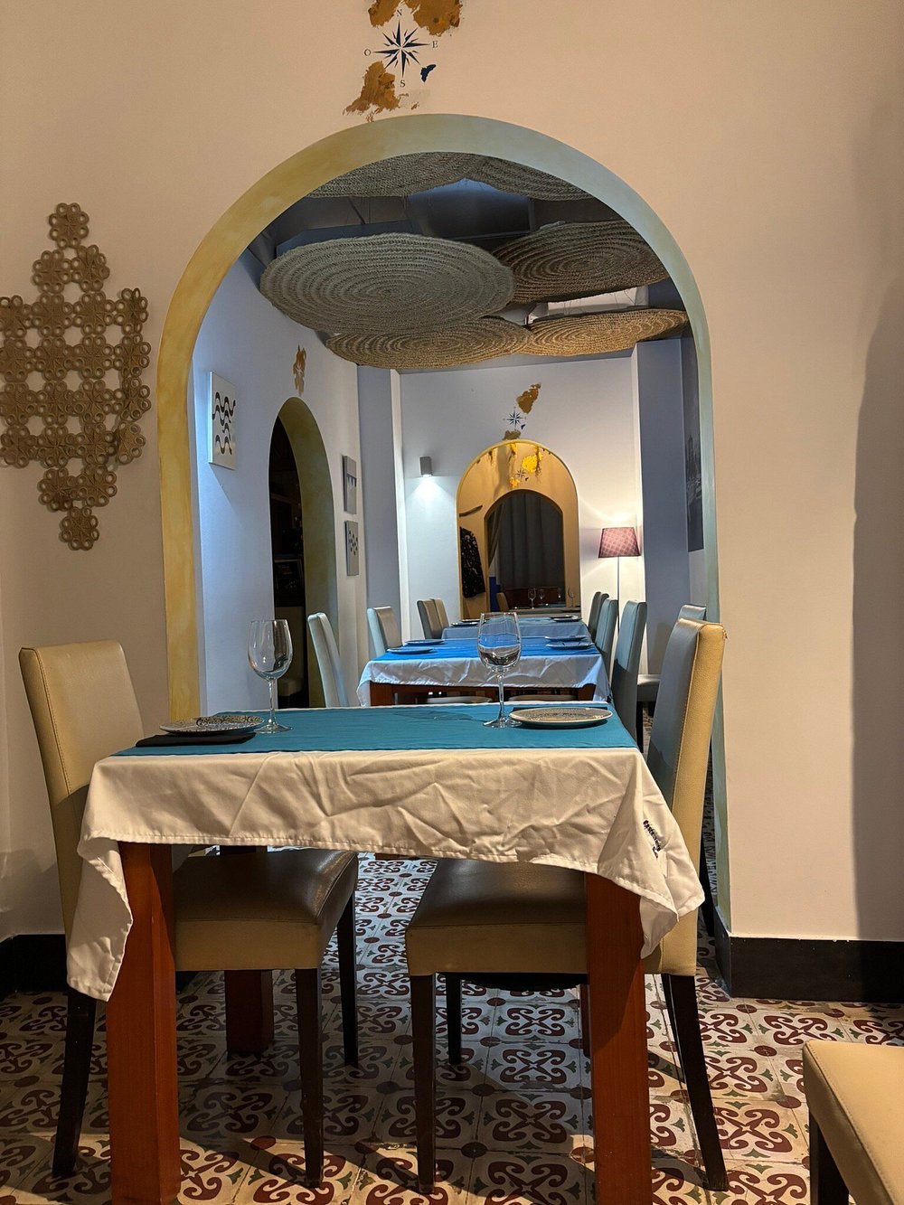 A dining room with a table set for four, featuring white tablecloths, wine glasses, and plates. The room has multiple arched doorways, wall decorations, and woven circular ceiling fixtures. The floor has patterned tiles.