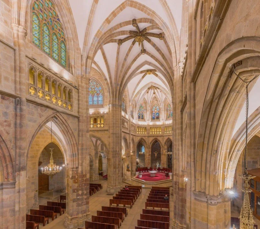 Interior of a Gothic cathedral with high vaulted ceilings, stained glass windows, and wooden pews arranged facing the altar at the front.