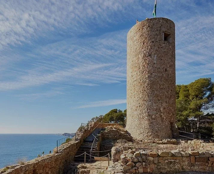 A tall, cylindrical stone tower with a small window near the top, overlooking the ocean with a blue sky and scattered clouds. There are stairs and a railing leading up to the tower, and a signal flag on a pole at the top. Trees are visible in the background.