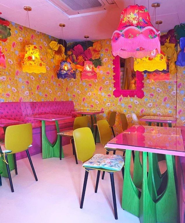 Colorful interior with yellow floral-patterned wallpaper, pink tufted seating, and bright multicolored hanging lamps.
