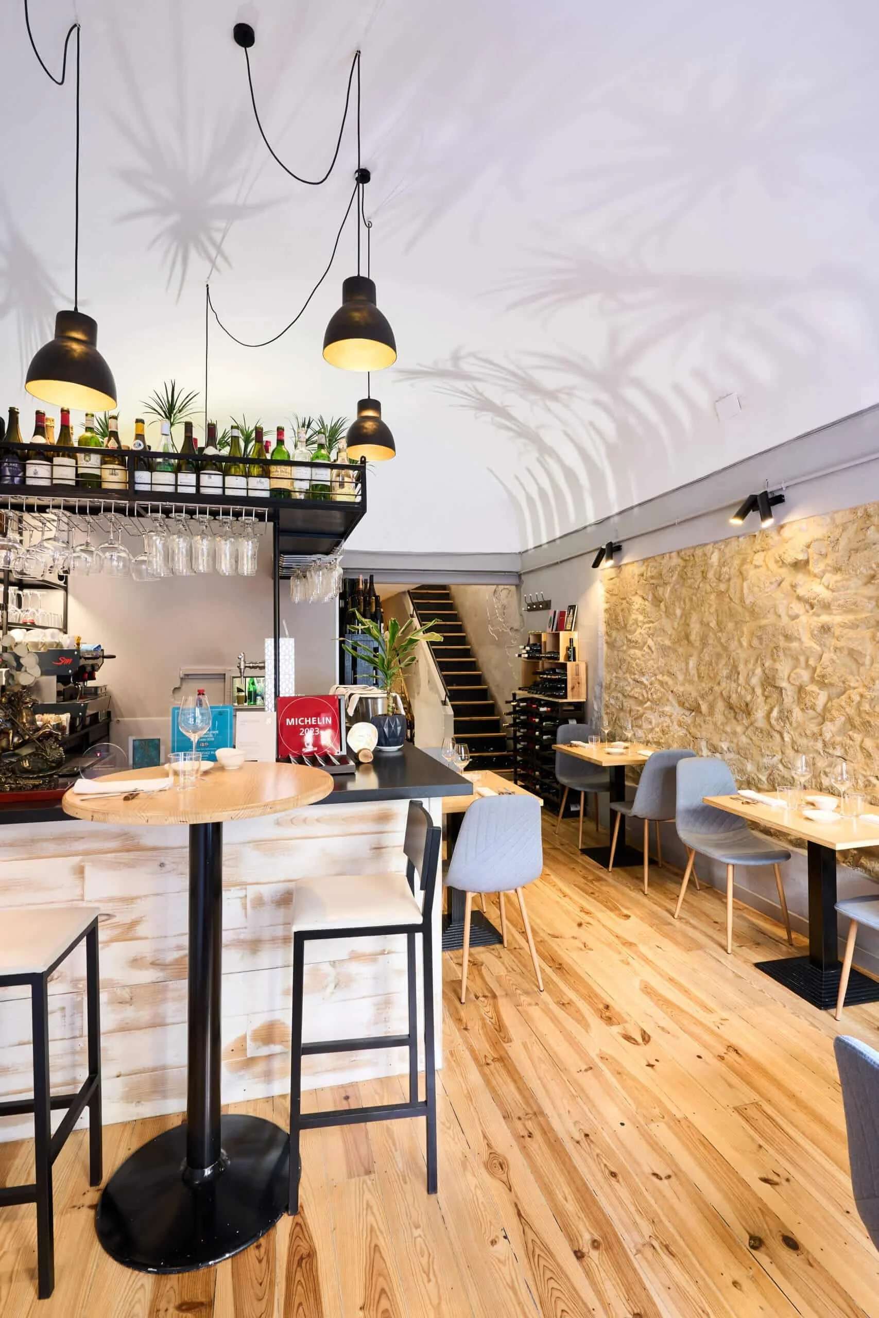 Interior of a modern restaurant with wooden floors, tables, chairs, a stone accent wall, and hanging black pendant lights. There is a bar area with bottles and glassware overhead. Shadows of palm leaves are cast on the ceiling.