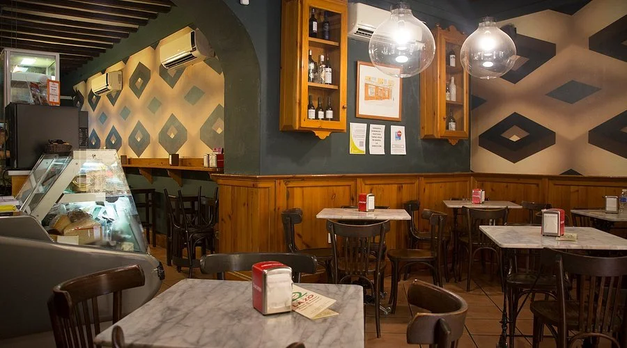 Interior of a cozy cafe with marble tables, wooden chairs, wall art, hanging glass lights, and a glass display case with desserts.