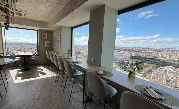 Modern dining area with a long breakfast bar by large floor-to-ceiling windows overlooking a cityscape with blue sky and clouds, decorated with plants and framed artwork.