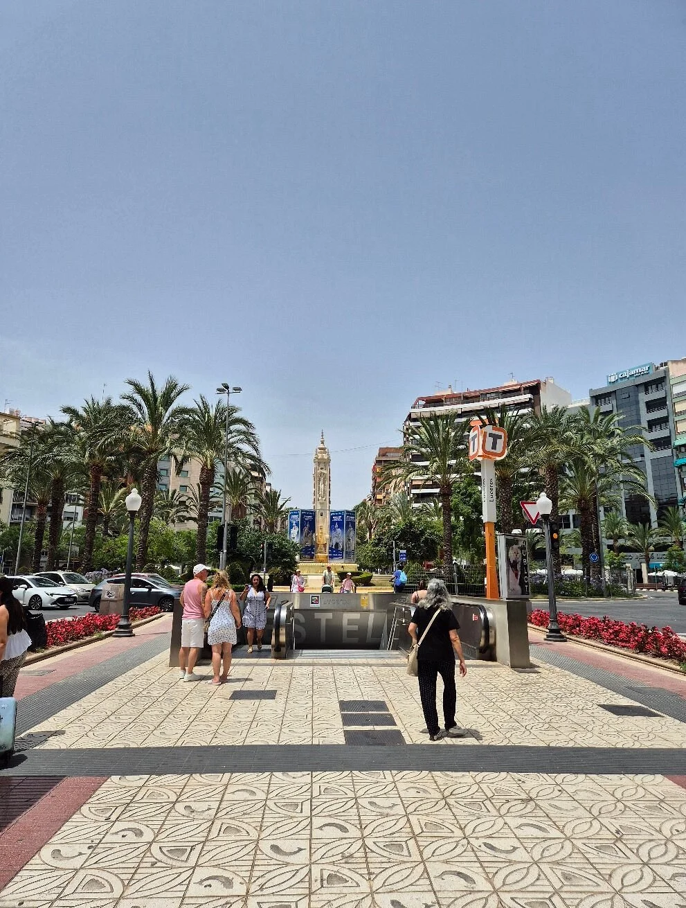 City square with palm trees, a fountain, and a tall clock tower in the background. People walking, some carrying luggage, and cars parked nearby.