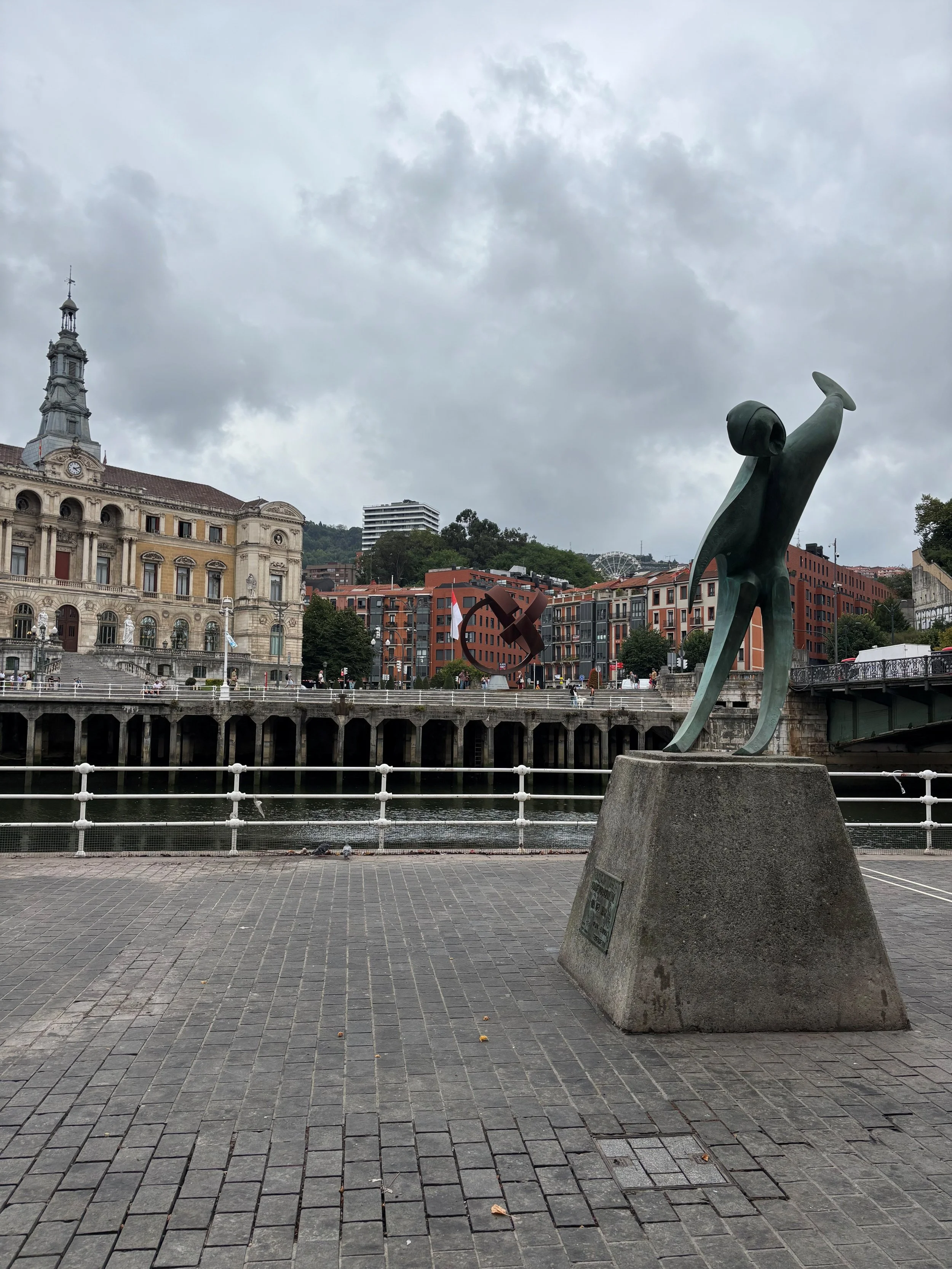A cityscape featuring historic and modern buildings with a statue in the foreground on a cloudy day.