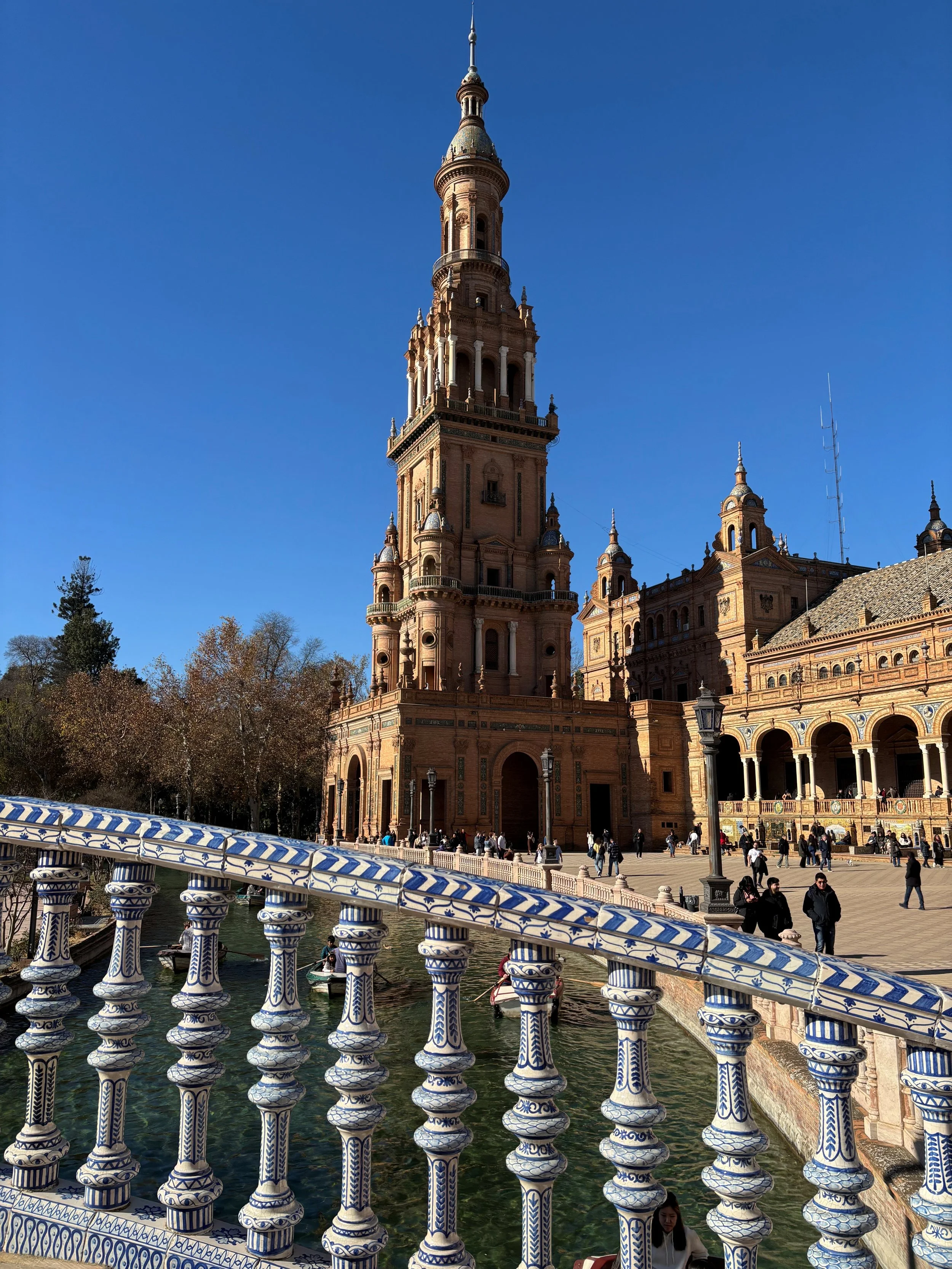 A large, ornate building with a tall tower, arched windows, and decorative details, situated next to a pond with a decorative white and blue railing, in a sunny setting with clear blue sky and people walking around.