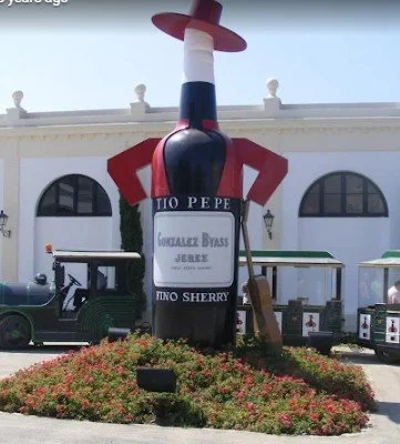 Large decorative bottle shaped like a condiment bottle, painted to resemble Ketchup, with a red cap and a label that reads 'TIO PEPE' and 'Gonzalez Byass Jerez.' It is displayed outside in front of a white building with arches and a flower bed surround.
