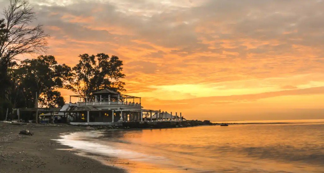 A beach at sunset with a pier and a building, calm water reflecting the orange sky, and trees in the background.
