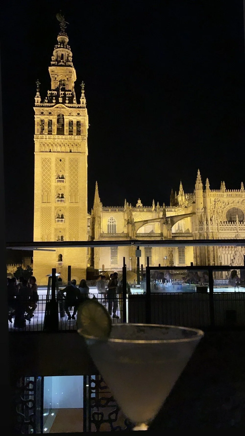 Night view of a historic cathedral with illuminated gothic architecture, including a tall clock tower, people sitting and standing on a terrace, and a large glass window in the foreground.
