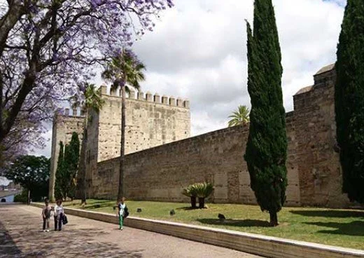Ancient stone fortress wall with a tower, tall cypress trees, and a few people walking nearby on a paved walkway under a cloudy sky.