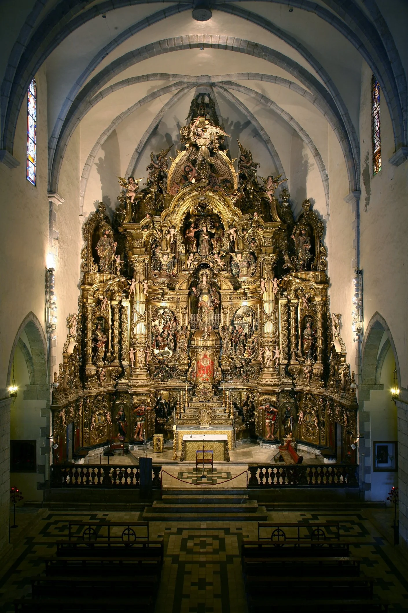 The interior of a historic church with an ornate gold altar featuring numerous religious sculptures and decorations, flanked by stained glass windows.