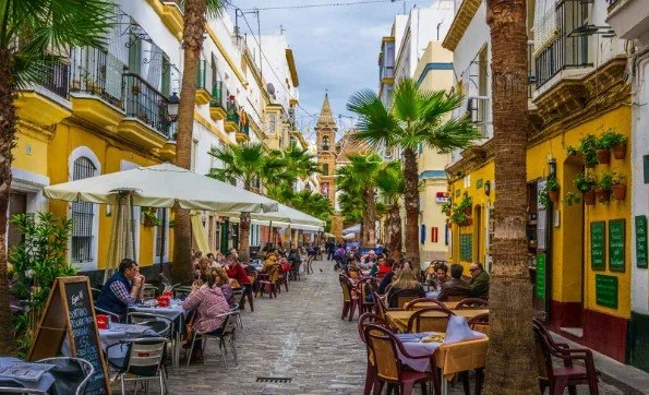 Outdoor dining street scene with tables, chairs, palm trees, and people dining under umbrellas in front of colorful buildings, with a church tower in the background.