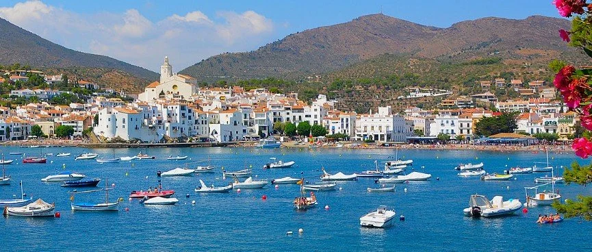 A coastal town with white buildings along the shoreline and boats in the water, mountains in the background, and pink flowers on the right side.