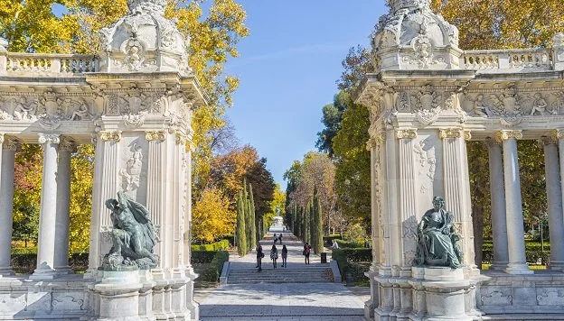 Entryway through ornate white stone gates into a park with walking paths, statues, and trees with autumn leaves.